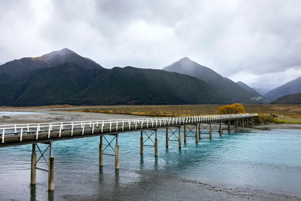 A long footbridge over a braided turquoise river under a cloudy mountain sky