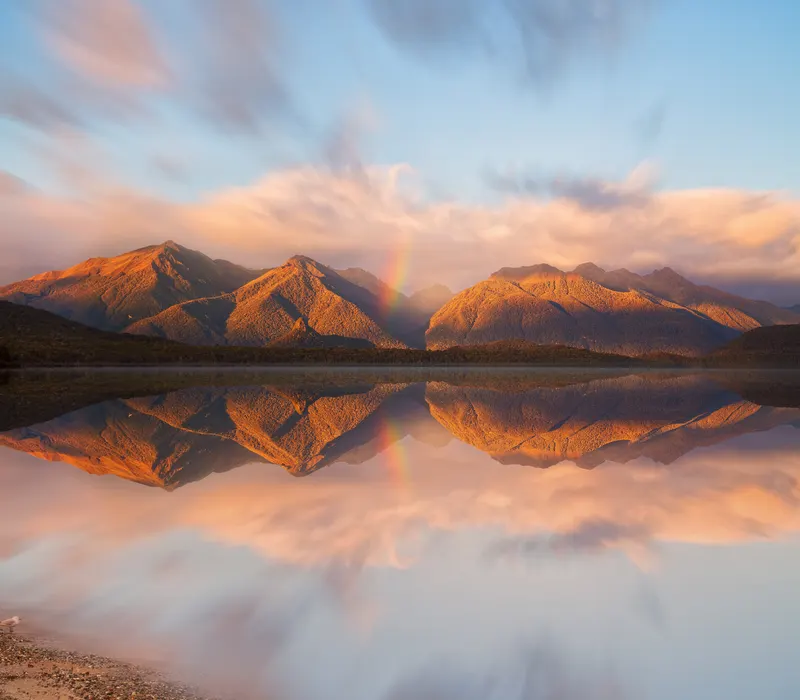 Dawn mountains and a rainbow reflected in still deep-south water